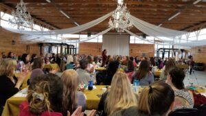 A large group of people is seated at tables covered with yellow tablecloths in a spacious hall with wooden walls and chandeliers. A person at the center is speaking, and the audience is attentively listening and applauding. White drapes hang from the ceiling.