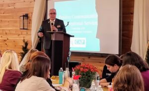A man in a suit and glasses speaks into a microphone at a podium in front of a projected screen. The screen displays "Positive Communication for Exceptional Services." Several attendees are seated at tables, some taking notes. The setting appears to be a seminar.