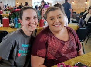 Two smiling women seated next to each other at an indoor event with tables and people in the background. One woman is wearing a gray t-shirt, and the other is in a maroon and pink plaid blouse. The setting features wooden walls and flower decorations on the tables.