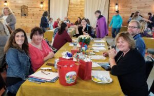 A group of people are sitting around a long table covered with a yellow tablecloth. The table has papers, snacks, and a red bucket filled with items. They are indoors, in a room with wooden walls. Some people are engaged in discussions.
