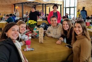 Five smiling women are sitting around a table with a Jenga game set up in the center. The setting is an indoor gathering with several other people in the background. Some autumn decorations and drinks are visible on the table, contributing to a cozy atmosphere.