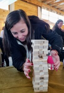 A woman with long brown hair and a black jacket carefully places a wooden block on a tall Jenga tower. The scene is set indoors in a wooden structure, with other people in the background. The tower is on a green surface.