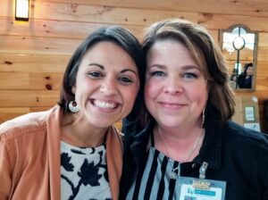 Two women are smiling and standing close together inside a wooden cabin-like venue. One woman, on the left, is wearing a coral blazer and the other woman, on the right, is wearing a name badge and a striped shirt. Both appear cheerful and friendly.