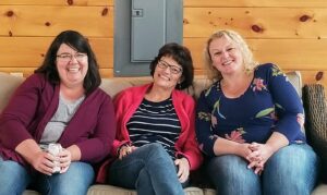 Three women sitting on a couch smiling and leaning towards each other. The woman on the left has dark hair, glasses, and is wearing a maroon sweater. The woman in the middle has short dark hair, glasses, and a red cardigan. The woman on the right has curly blonde hair and a floral top. The background is a wooden wall.