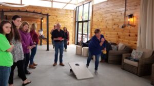A group of people is gathered indoors, playing a game of cornhole. One woman is mid-throw, aiming at the cornhole board in front of her. Others are standing around, watching the game. The room has wooden walls, large windows, and some seating arrangements.