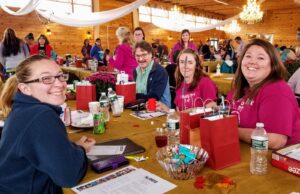 A group of four people sits at a decorated table during an indoor event. Three women wear matching pink shirts and a man in glasses and a green shirt smiles at the camera. Party favor bags, snacks, and drinks are on the table. Other attendees are in the background.