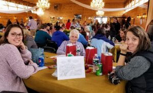 A group of three women seated at a table in a busy, warmly lit hall. The table has coloring activities, soda cans, gift bags, and snacks. Other tables with people are in the background, and two chandeliers hang from the wooden ceiling.