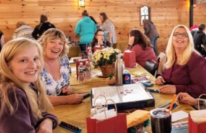 A group of people sitting around a table with food, drinks, and board games on it. Three women in the foreground are smiling at the camera. The background shows more people engaging in activities inside a warmly lit, wood-paneled room.