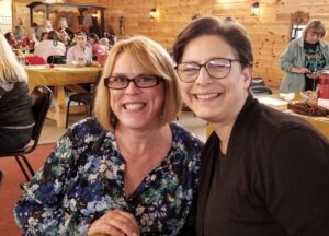 Two women are smiling and posing for a photo indoors. They are seated in a rustic room with wooden walls and tables adorned with tablecloths. Other people are seated in the background, chatting and enjoying a meal. The atmosphere is warm and convivial.