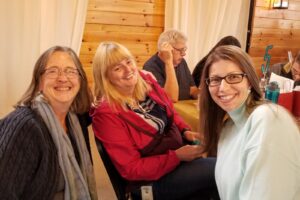 Three women are sitting and smiling at the camera in a cozy setting with wooden walls. The women are dressed warmly; one wears a gray sweater, another a red jacket, and the third a light blue sweater. In the background, there are other people and a numbered table.