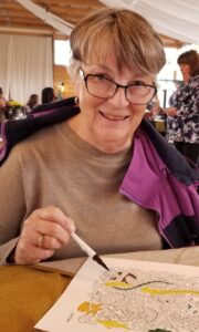 An older woman with short gray hair and glasses is smiling while holding a marker and coloring a detailed drawing. She is wearing a beige sweater with a purple jacket draped over her shoulders. She is seated indoors at a table with people in the background.