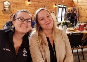 Two women sitting and smiling in a wooden cabin setting. The woman on the left, wearing glasses and a dark jacket labeled "Kristina" and "MG", has her arm around the woman on the right, who has blonde hair and is wearing a beige cardigan. People are gathered in the background.