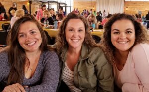 Three women are sitting close together at a social event, smiling at the camera. They are seated at a table in a spacious indoor area with wooden walls and other people in the background engaged in various activities.