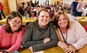 Three smiling women are seated at a table in a bustling indoor setting with many people in the background. The woman on the left is wearing glasses and a pink top, the middle woman is in a gray sweater, and the woman on the right is wearing a light pink cardigan.