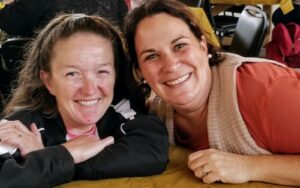Two women are smiling as they lean their heads on their hands across a table. One wears a black jacket, and the other is dressed in a maroon top and beige vest. Both appear to be in a cheerful, casual setting.