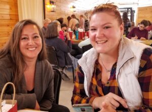 Two women are sitting at a table in a cozy, wood-paneled room filled with people. Both are smiling at the camera. One woman has long brown hair and is wearing a dark sweater, while the other has her hair tied back and is wearing a checkered shirt and a white vest.