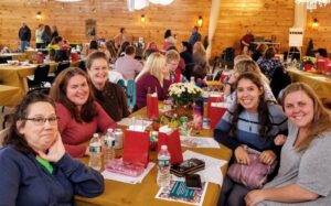 A group of six women is seated around a decorated table at a social event in a wooden-paneled hall. They are smiling and chatting, with various items, drinks, and gift bags on the table. In the background, more people are mingling and sitting at other tables.