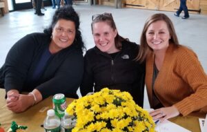 Three women are sitting at a table decorated with a pot of yellow flowers. They are smiling at the camera. The woman on the left wears a black top, the woman in the middle has a black jacket, and the woman on the right is in an orange sweater. Drinks are on the table.