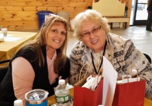 Two women smiling and sitting at a table with gift bags and a water bottle. One woman is wearing a black vest over a pink top, and the other is wearing a patterned sweater with glasses. The setting appears to be indoors with wooden walls and tables in the background.