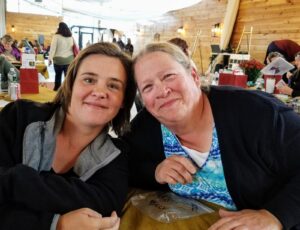 Two women seated at a table in a bustling wooden event hall, smiling warmly at the camera. The woman on the left has short dark hair and is wearing a grey jacket, while the woman on the right has long hair tied back and is wearing a blue patterned shirt.