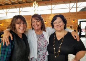 Three women stand together indoors, smiling warmly at the camera. The woman on the left wears rimmed glasses, a blue plaid shirt, and a black vest. The middle woman wears a patterned blouse and has a hand on each of the other women's shoulders. The woman on the right wears a black top and holds a coffee cup. The room has wooden walls and a chandelier above.