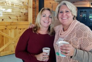 Two women are smiling at the camera while holding disposable coffee cups. They are indoors in a wooden room with one woman wearing a red sweater and the other in a cream-colored knit sweater with a scarf. The background features a wooden door with a black frame.