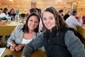 Two women are seated at a wooden table, smiling at the camera. They are in a warm, indoor setting with wooden walls and other people sitting at tables in the background. The table has papers, pens, and a water bottle on it.
