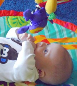 A baby lies on a colorful play mat, gazing up at a purple stuffed animal hanging from an activity gym. The baby is wearing a white onesie with a dark bear design and has their hands near their mouth. Bright hues of green, red, and blue surround the baby.