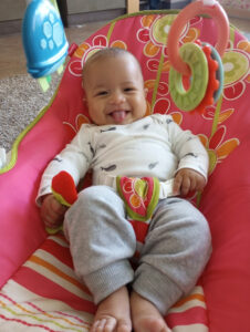 A baby in a white shirt with gray pants sits in a colorful pink and red baby chair, smiling with their tongue out. They are holding a toy and have other toys hanging above them. The baby seems happy and content.