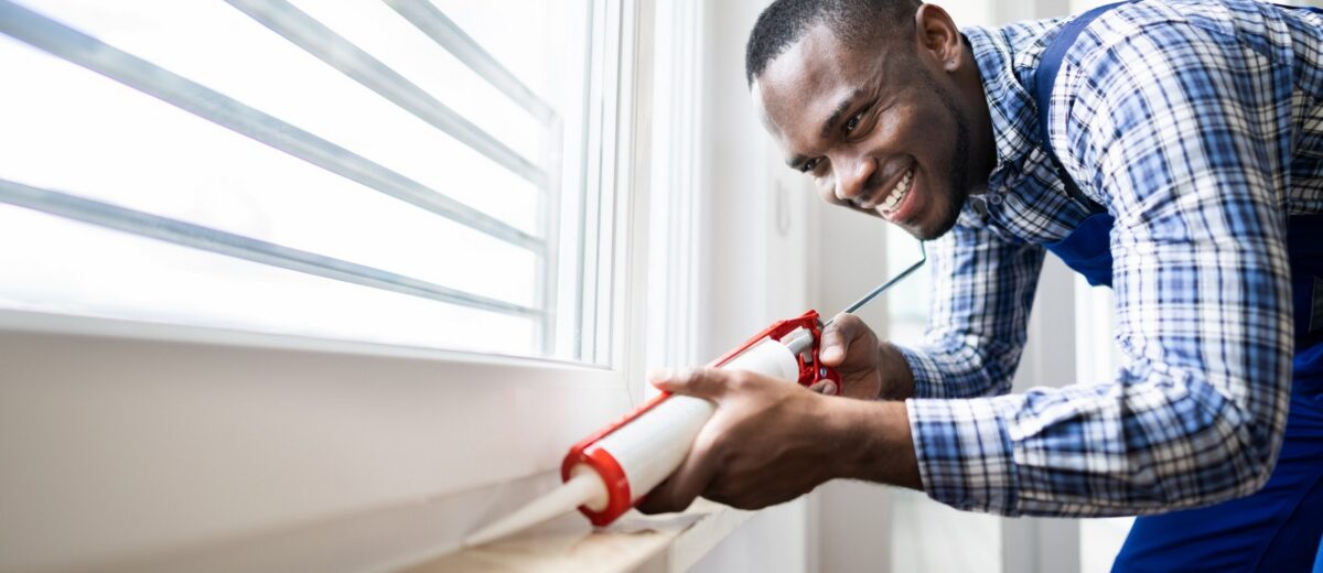 A man in a checkered shirt and blue overalls is smiling while using a caulking gun to seal the inside frame of a window. Bright natural light illuminates his workspace, which appears to be part of a modern home.