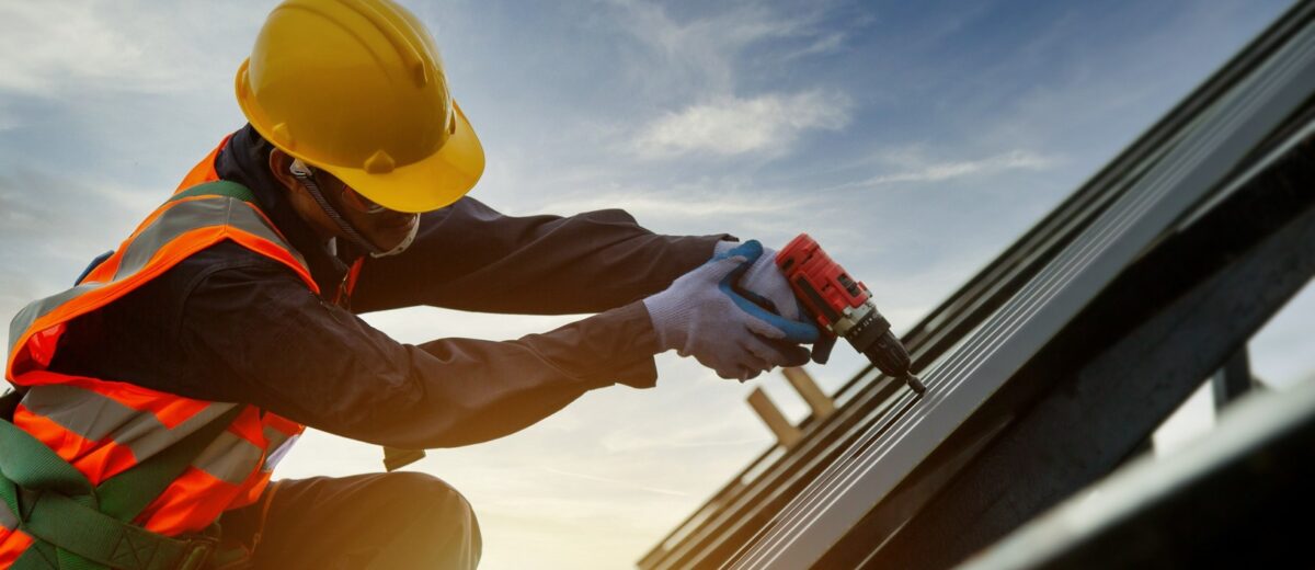 A construction worker wearing a yellow hard hat, orange reflective vest, and safety gloves is using a power drill to install solar panels on a roof. The sky is clear with a few clouds, creating a bright and productive work environment.