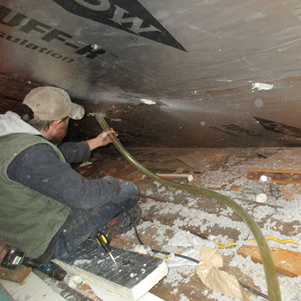 A person wearing a cap and work clothes is crouched in an attic space, using a hose for insulation. The floor is covered with insulation material, tools, and debris. An insulation board is partially installed above the person.