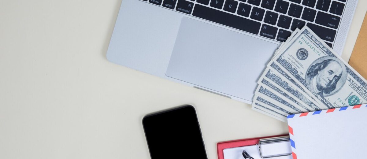 A laptop, a stack of hundred-dollar bills, a smartphone, an envelope, a pair of glasses, and a small clipboard are arranged on a white surface.
