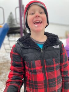 A child wearing a red and black plaid jacket and a matching hat is standing outside near playground equipment. The ground appears snowy, and the child is smiling with snow on their face. The background shows a snowy playground scene with another child partially visible.