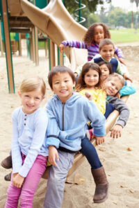 A group of seven children smiling and sitting in a line on a playground slide. They appear to be of diverse ethnic backgrounds. The playground is outdoor with a sandy ground, and trees are visible in the background. The children are dressed in casual clothing.